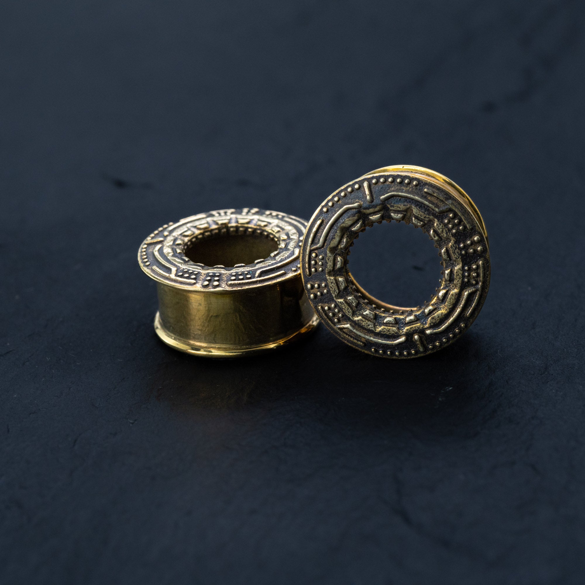 Close-up of two Kaya Movement's TERA Futuristic Sci Fi Flesh Tunnels in Gold Brass against a dark background. One gauge stands upright showcasing its intricate, circular Aztec-style design, while the other lies on its side revealing its hollow center, embodying the elegance and innovation of futuristic ear tunnels available in sizes ranging from 8mm to 20mm.